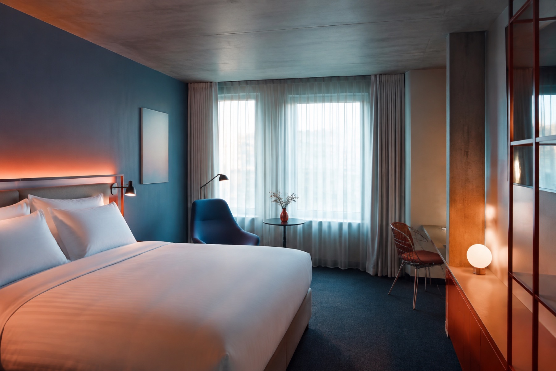 Hotelkamer met blauwe wand, wit opgemaakt bed en een oranje tafellamp